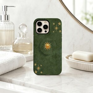 May include: A dark green smartphone case featuring a gold sun emblem in the centre and gold star motifs scattered across the top and bottom corners. The case includes a circular grip attachment on the back and a cutout for the camera lens.