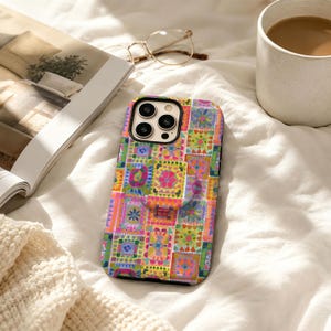 Könnte beinhalten: Eine Smartphone-Hülle mit einem lebendigen, mehrfarbigen Patchwork-Muster aus kleinen Quadraten mit verschiedenen Blumen- und geometrischen Motiven in Rosa, Orange, Grün, Blau und Gelb, inklusive eines runden, ausklappbaren Griffs auf der Rückseite.
