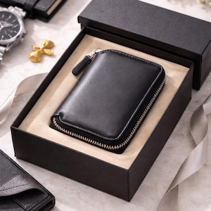 May include: A black leather wallet with a gold zip is presented in a black gift box. The wallet is rectangular and has a zip closure. The box is lined with beige material. Other items in the image include a watch, pen, and notebook.