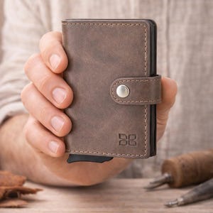 May include: A brown leather wallet with a snap closure and a small logo. The wallet is held in a person's hand. The wallet is rectangular and appears to be made of textured leather.