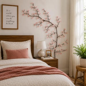 Op de afbeelding: Een slaapkamer met een houten bedframe, een crèmekleurig dekbed en een roze deken. Een ingelijste print met de tekst "In a world where you can be anything, be kind" hangt boven het bed. Een decoratieve kersenbloesemtak siert de muur.