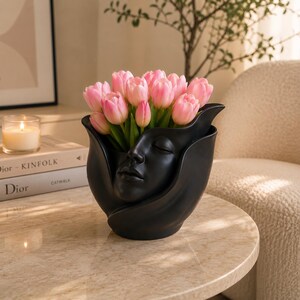 May include: A black ceramic vase with a sculpted face design, holding a bouquet of pink tulips. The vase has a unique shape, with the face integrated into the design. The tulips are a vibrant pink, creating a striking contrast against the dark vase.