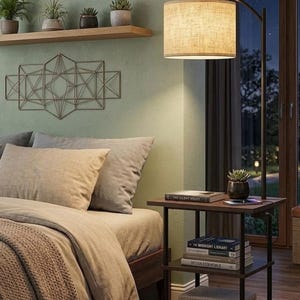 May include: A bedroom scene featuring a bed with beige bedding and a brown throw blanket. A dark brown side table with books and a small plant is next to the bed. A floor lamp with a beige lampshade illuminates the room. Decorative wall art and potted plants are also visible.