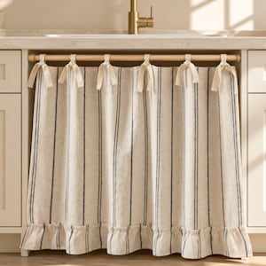 May include: A beige kitchen sink curtain featuring vertical dark gray stripes and a ruffled bottom hem. The curtain is suspended from a light-colored wooden rod by six fabric ties knotted at the top, creating a gathered, pleated appearance.