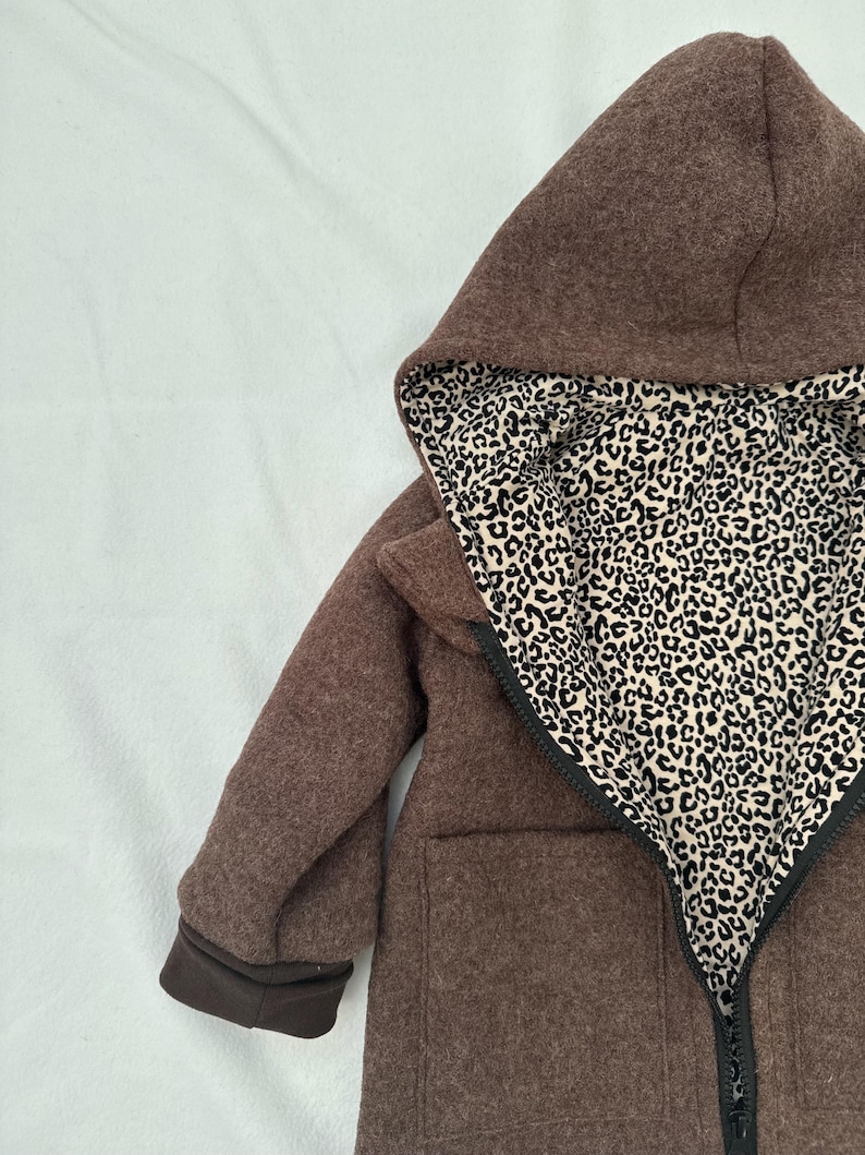 May include: A brown hooded jacket featuring a black zipper closure and a contrasting interior lining with a black and beige leopard print pattern. The garment includes long sleeves with dark brown cuffs and two front pockets on the exterior.