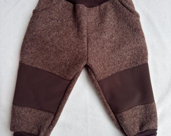Puede incluir: Pantalones marrones de tela texturizada similar a la lana, con una cintura elástica ancha de color marrón oscuro y puños a juego en los tobillos. Las piernas incluyen paneles lisos de color marrón oscuro en las rodillas que contrastan con el tejido.