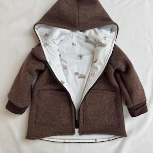 May include: A brown hooded jacket with a white interior lining featuring a small, colorful mountain and floral pattern. The jacket has a black front zipper, long sleeves with dark brown cuffs, and two square front pockets on the outer shell.