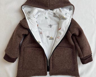May include: A brown hooded jacket with a black front zip and two front pockets. The interior lining is white and features a repeating pattern of small grey mountains, brown leaves, and floral shapes. The sleeves have dark brown ribbed cuffs.