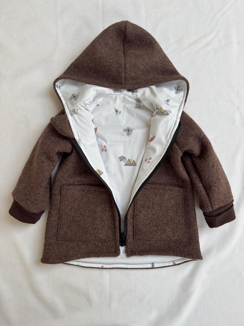 May include: A brown hooded jacket with a white interior lining featuring a small, colorful mountain and floral pattern. The jacket has a black front zipper, long sleeves with dark brown cuffs, and two square front pockets on the outer shell.