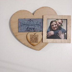 May include: Wooden heart-shaped photo frame with a small square frame and a heart-shaped QR code. The frame has a denim-like patch with the word "love" stitched across it. The square frame holds a photo of a couple.
