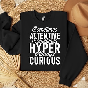 Op de afbeelding: Zwarte crewneck sweatshirt met witte tekst: "Sometimes ATTENTIVE Sometimes HYPER Always CURIOUS". De sweatshirt wordt weergegeven op een geweven oppervlak met een bruine hoed op de achtergrond.