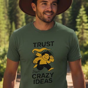 Könnte beinhalten: Olivgrünes T-Shirt mit dem Text "TRUST your CRAZY IDEAS" und einer Cartoon-Grafik. Die Person trägt einen braunen Hut. Der Hintergrund zeigt ein Gebirge und einen Weg durch einen Wald.