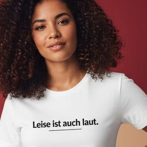 Könnte beinhalten: Ein weißes T-Shirt mit Rundhalsausschnitt und dem deutschen Text "Leise ist auch laut" in Schwarz, unterstrichen. Das Model trägt eine blaue Jeans und hat lockiges braunes Haar. Der Hintergrund ist eine Mischung aus Rot und Beige.