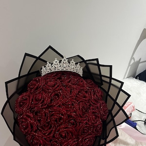 May include: A bouquet of deep red glittery roses wrapped in black paper, topped with a silver-toned metal tiara featuring clear crystal accents. The roses are arranged in a dense, rounded cluster, and the tiara sits prominently on the top centre.