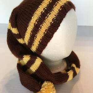May include: A brown and yellow striped knit hat with a long tail and a yellow pom-pom. The hat is designed to fit over the head and has a long, scarf-like extension. The hat is displayed on a white mannequin head.