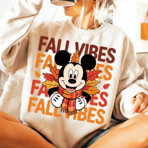 Può includere: Una felpa girocollo color crema con una grafica di Topolino che indossa una sciarpa a quadri, circondato da foglie autunnali colorate. Il testo "FALL VIBES" è ripetuto in lettere marroni, arancioni e rosse sulla parte anteriore del capo.