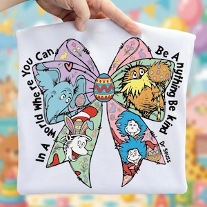 May include: White t-shirt featuring a colorful bow design with Dr. Seuss characters and Easter eggs. The bow includes the text "In A World Where You Can Be Anything Be Kind". The design incorporates various pastel colors and cartoon illustrations.