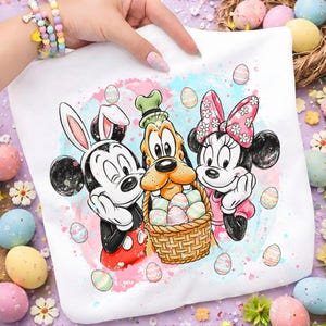 May include: White t-shirt featuring Mickey Mouse with bunny ears, Goofy, and Minnie Mouse, holding a basket of Easter eggs. The design has a watercolor background with pastel colors and Easter egg accents.