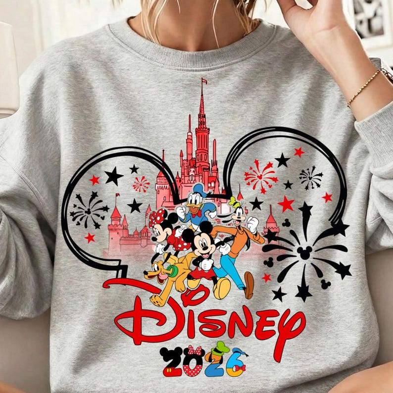 K&ouml;nnte beinhalten: Graues Sweatshirt mit einem Disney-Design. Das Design zeigt Micky Maus, Minnie Maus, Donald Duck, Goofy und Pluto, mit einem Schloss und Feuerwerk. Das Wort "Disney" ist rot, und "2026" ist in bunten Zahlen.