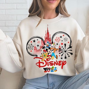 K&ouml;nnte beinhalten: Cremefarbenes Sweatshirt mit Disney-Design. Das Design zeigt Micky Maus, Minnie Maus, Donald Duck, Goofy und andere Charaktere vor einem Schloss. Das Wort "Disney" ist rot, darunter steht "2026".