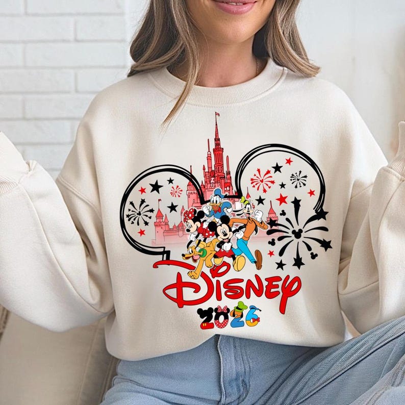 K&ouml;nnte beinhalten: Cremefarbenes Sweatshirt mit Disney-Design. Das Design zeigt Micky Maus, Minnie Maus, Donald Duck, Goofy und andere Charaktere vor einem Schloss. Das Wort "Disney" ist rot, darunter steht "2026".