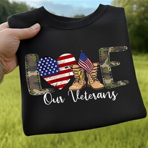 Pode incluir: Camisola preta com a palavra "LOVE" num padrão de camuflagem. O "O" é um coração com o design da bandeira americana. A imagem inclui botas de combate, uma bandeira americana e o texto "Our Veterans."