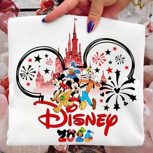 K&ouml;nnte beinhalten: Wei&szlig;es T-Shirt mit einem farbenfrohen Disney-Design. Das Design zeigt Micky Maus, Minnie Maus, Donald Duck, Goofy und Pluto, ein rotes Schloss, Feuerwerk und das Wort "Disney" in Rot. Das Jahr "2026" ist ebenfalls enthalten.