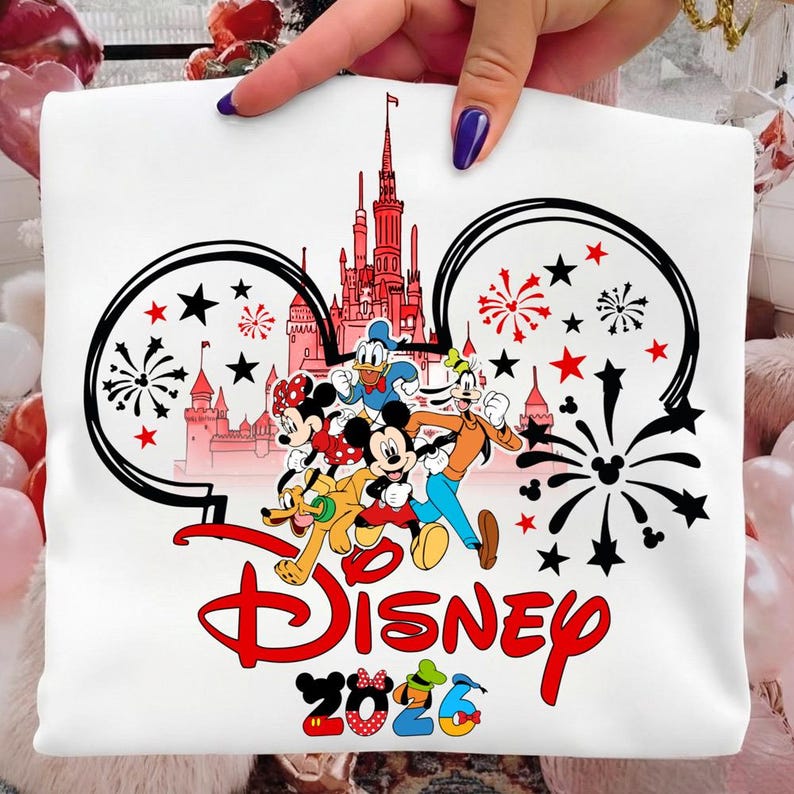 K&ouml;nnte beinhalten: Wei&szlig;es T-Shirt mit einem farbenfrohen Disney-Design. Das Design zeigt Micky Maus, Minnie Maus, Donald Duck, Goofy und Pluto, ein rotes Schloss, Feuerwerk und das Wort "Disney" in Rot. Das Jahr "2026" ist ebenfalls enthalten.