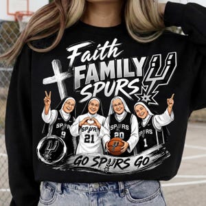 May include: A black sweatshirt featuring a graphic of four nuns wearing San Antonio Spurs basketball jerseys. The text reads Faith, FAMILY, SPURS, and GO SPURS GO. The design includes a cross, a basketball, and the Spurs team logo in white and black.