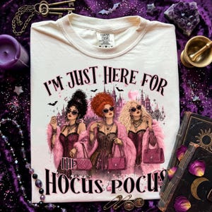 Könnte beinhalten: Ein cremefarbenes T-Shirt mit einer Grafik von drei stilisierten Figuren mit dunklem, rotem und blondem Haar, die rosa Pelzmäntel und Sonnenbrillen tragen. Der Text I'M JUST HERE FOR THE HOCUS POCUS steht über und unter den Figuren vor einem Schloss.