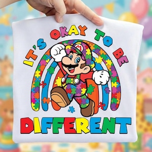 Peut inclure: T-shirt blanc avec un personnage de dessin animé en tenue rouge et verte, un arc-en-ciel fait de pièces de puzzle et le texte "It's Okay To Be Different". Le design est vif et coloré.