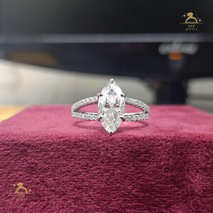 May include: A marquise-cut diamond engagement ring with a split shank band. The ring features a large, pear-shaped center stone and a band adorned with smaller, pavé-set diamonds. The ring is set on a burgundy velvet surface.