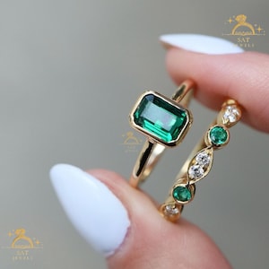 May include: Two gold rings. One ring features a rectangular emerald-cut green gemstone. The other ring has three round green gemstones and three small diamonds. The rings are held up by a person.