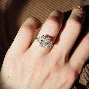 Puede incluir: Un anillo de oro con un diamante grande de talla rectangular flanqueado por dos diamantes más pequeños de talla cuadrada. El anillo está engastado en una fina banda de oro, resaltando el brillo de las piedras preciosas. El anillo se muestra en una mano.