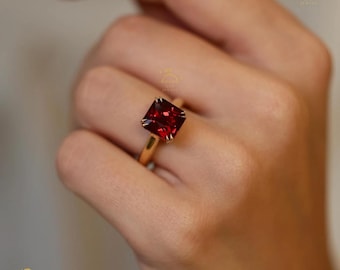 以下が含まれることがあります： 正方形にカットされた、深い赤色の宝石をあしらったゴールドリング。 宝石は4本爪のセッティングで、バンドはシンプルなポリッシュゴールドです。 リングは指に装着されており、石の鮮やかな色を際立たせています。