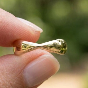 May include: A small, polished gold-toned ring featuring a smooth, hourglass-shaped design that tapers in the center. The metal surface is highly reflective, catching the light to create bright highlights along its curved, rounded edges.