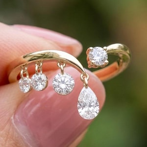 May include: A gold ring with a distinctive design, showcasing several dangling diamonds. The ring has a curved band with a large diamond at the top and four smaller diamonds hanging below, including a pear-shaped diamond. The ring is held by a person.
