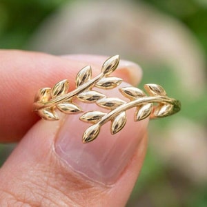May include: A gold ring featuring a design of two overlapping branches with small, smooth leaves. The metal has a polished, reflective finish, and the band is shaped to mimic the organic, curved structure of natural foliage stems.