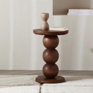 May include: A dark brown wooden side table with a unique design. The table features a round top, supported by three stacked spherical shapes, and a round base. A beige vase and a book rest on top. The table is approximately 24 inches tall.