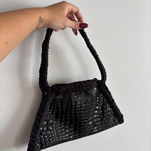 Peut inclure: Un sac à main noir doté d'une finition brillante et texturée façon croco sur le corps principal. Le sac est bordé d'une épaisse garniture en tissu noir tressé qui se prolonge en une anse assortie, créant un contraste de texture marqué.