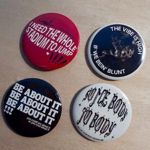 May include: Four circular pin-back badges featuring distinct designs. One is red with white text reading I NEED THE WHOLE STADIUM TO JUMP. One is black with white text THE VIBE IS HIGH IF WE BEIN' BLUNT. One is black with white text BE ABOUT IT. One is white with black text SOME BODY TO BODY.