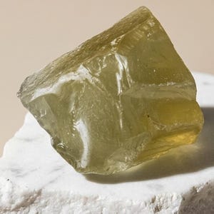 Puede incluir: Un trozo de vidrio crudo translúcido de color verde oliva pálido con bordes irregulares y facetados y una superficie lisa y reflectante, que descansa sobre una losa de mármol blanco dentada con textura rugosa y vetas grises.