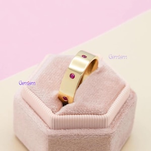 May include: A gold band ring featuring three small, round, flush-set pink gemstones spaced evenly along the outer surface. The ring is positioned within a soft, light pink velvet jewelry box. The word Gemism is printed in purple text.