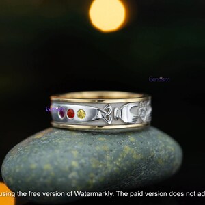 May include: A two-tone metal ring featuring a silver-colored band with gold-colored edges. The band is inlaid with three small round gemstones in red, orange, and yellow, followed by a silver-colored Celtic knot and a Claddagh symbol design.