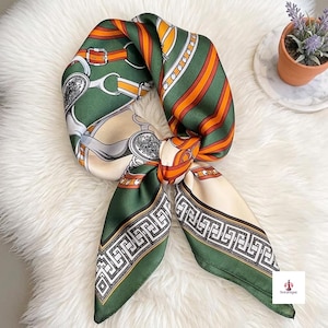 May include: A square scarf featuring a dark green base with orange and white diagonal stripes, equestrian-inspired buckle and harness motifs, and a black and white Greek key border pattern. The fabric has a smooth, satin-like finish.