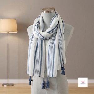 Puede incluir: Una bufanda blanca con rayas verticales azules de distintos anchos. La bufanda está colocada sobre un maniquí y rematada con pequeñas borlas azules en el dobladillo inferior. El texto Soravique es visible en la esquina inferior derecha.