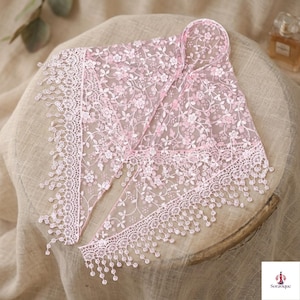 May include: A triangular pink lace head covering featuring an intricate floral pattern and delicate scalloped edges with dangling lace fringe. The sheer fabric displays a repeating botanical motif throughout the entire piece.