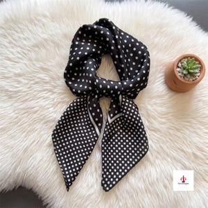 May include: A square scarf featuring a black background with a repeating white polka dot pattern. The scarf is folded into a knot shape, displaying two pointed ends. A small square logo with the text Soravique is visible in the bottom corner.