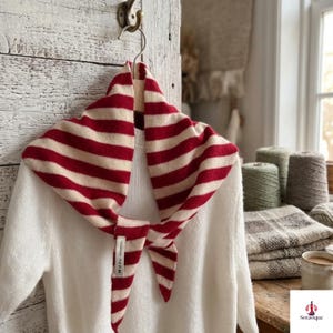 May include: A triangular scarf featuring horizontal red and cream stripes, tied at the front. The scarf is draped over a white knit sweater and includes a small white fabric tag with the text Soravique printed in black near the knot.