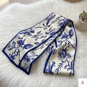 May include: A long, rectangular scarf featuring a cream-colored base with a repeating dark blue floral and vine pattern. The scarf has a solid dark blue border along its edges, creating a distinct contrast against the intricate botanical print.